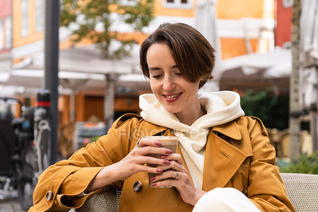 Donna positiva in capispalla con caffè caldo seduto a tavola in street cafè guardando in basso — Foto stock