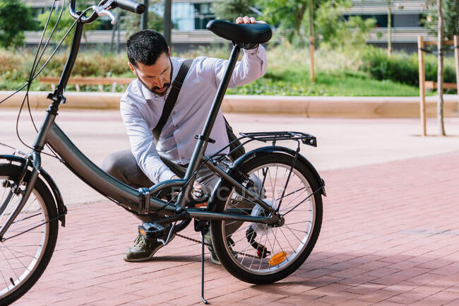 Voller Körper eines erwachsenen bärtigen Mannes in offizieller Kleidung, der die Rückenkette eines Fahrrads im Park im Zentrum des Bezirks fixiert — Stockfoto