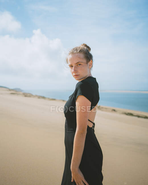Vista laterale di una femmina in abito scuro passeggiando su dune sabbiose nel deserto e sullo sfondo il mare guardando la macchina fotografica — Foto stock