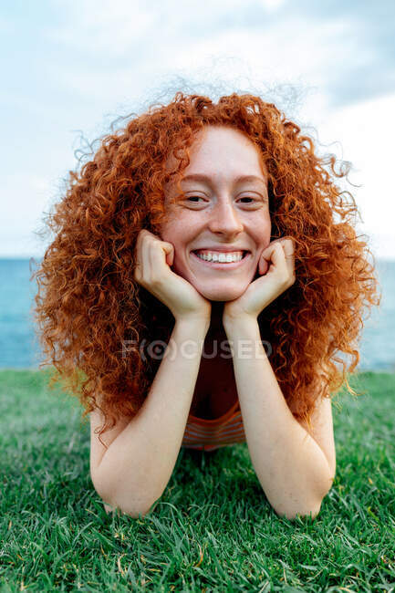 Happy curly redhead haired female with freckles lying on lawn looking at camera on coast of sea — Stock Photo