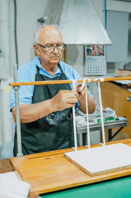 Attentive senior male artisan in apron and eyeglasses tying tapes on wooden board before working on printing press machine — Stock Photo