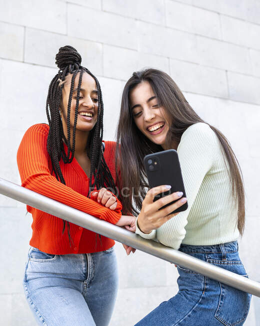 Снизу веселые разнообразные женщины в стильной одежде, стоящие на лестнице возле перила и смеющиеся во время просмотра смартфона на улице в дневное время — стоковое фото