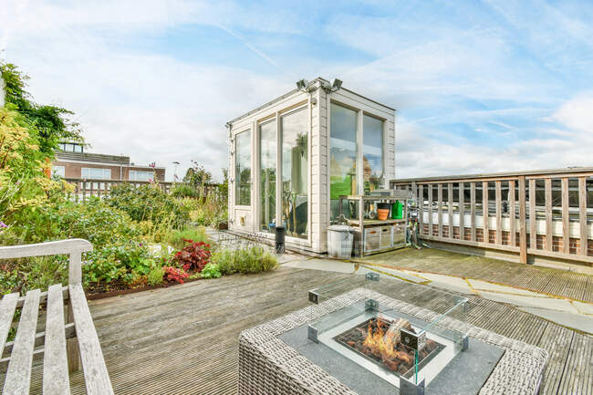 Holzmöbel und Glaskamin auf Rattan-Korbtisch auf dem Dach des modernen Gebäudes mit frischen grünen Pflanzen gegen wolkenverhangenen blauen Himmel am sonnigen Tag dekoriert — Stockfoto