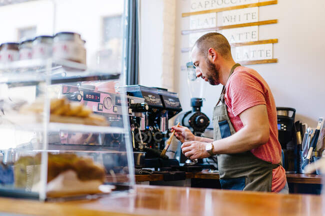 Side view of focused male barista in apron cleaning coffee machine with brush while standing near counter with desserts in coffee house — Stock Photo