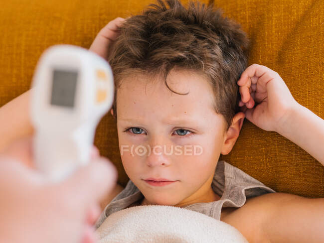 Dall'alto di genitore di coltura con termometro infrarosso che misura la temperatura di bambino malato triste che si trova sotto coperta sul divano a casa — Foto stock