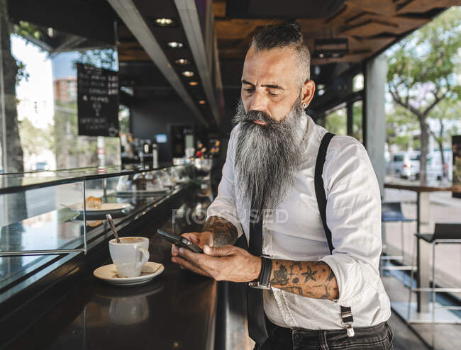 Concentrated male hipster in formal wear text messaging on cellphone while standing near counter with cup of coffee in cafeteria — Stock Photo