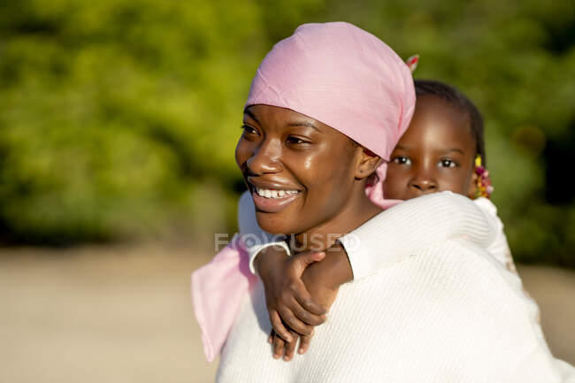 Seitenansicht einer positiven schwarzen Frau mit rosa Kopftuch, die bei sonnigem Wetter ihre kleine Tochter auf dem Rücken gegen verschwommene grüne Bäume trägt — Stockfoto