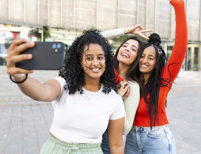 Fröhliche junge multiethnische Freundinnen in trendigen Outfits stehen mit erhobenen Armen und lächeln, während sie bei Tageslicht ein Selfie mit dem Smartphone auf der Straße machen — Stockfoto