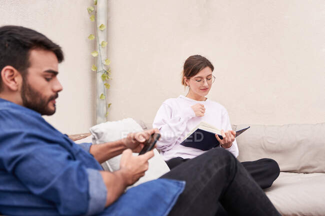 Concentrated bearded male surfing social media on cellphone while sitting on couch near friend reading book in spare time — Stock Photo