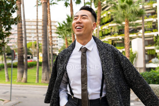 Добре одягнений молодий веселий азіатський чоловічий підприємець у краватці дивлячись у далечінь, коли крокує по дорозі проти сучасних будинків у місті — стокове фото