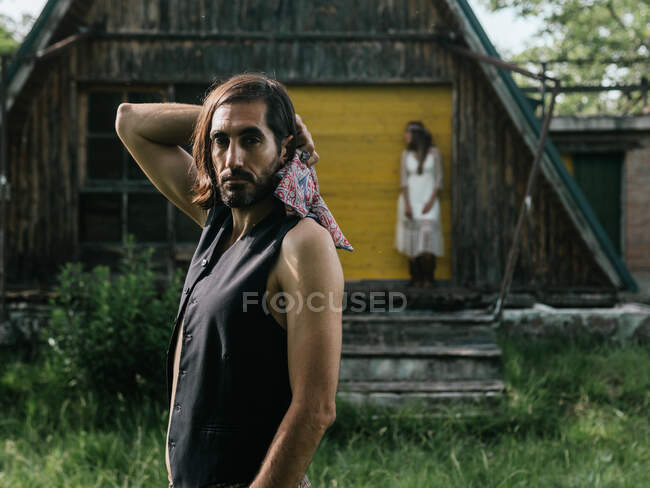 Close-up de um homem sério olhando para a câmera e no fundo uma menina borrada encostada a uma casa de madeira abandonada — Fotografia de Stock
