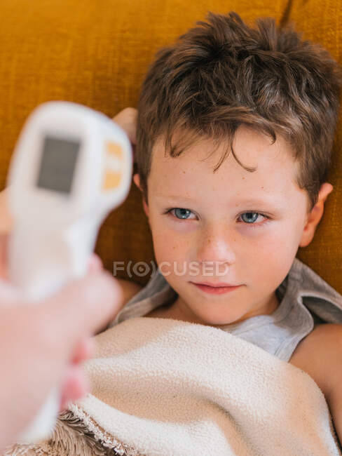 Von oben ein Elternteil mit Infrarot-Thermometer, das die Temperatur eines traurigen kranken Kindes misst, das zu Hause unter einer Decke auf dem Sofa liegt — Stockfoto