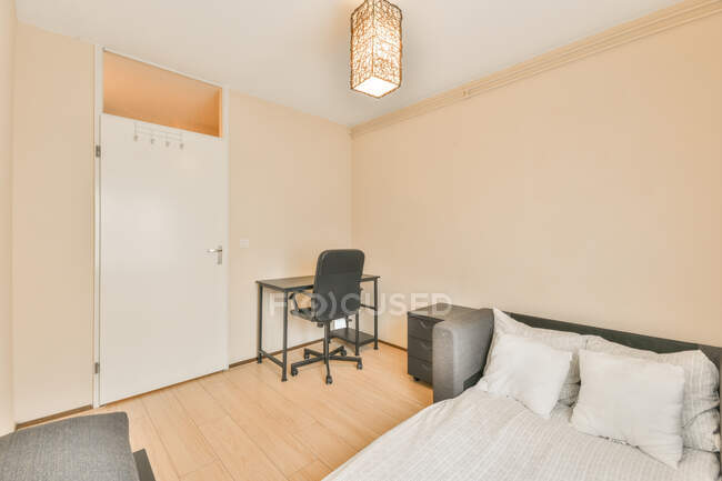 Interior del dormitorio con diseño moderno con paredes de color beige y puerta blanca dotada de sofá-cama hecha con una cubierta ligera y cojines colocados cerca de una pequeña mesita de noche y sillón iluminado con lámpara decorativa - foto de stock