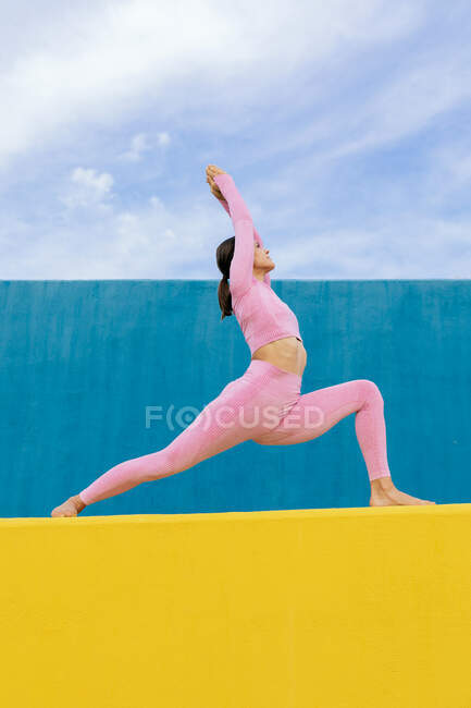 Von unten eine junge Frau in Sportkleidung, die in Virabhadrasana an einer gelben Wand vor blauem Hintergrund und bewölktem Himmel steht — Stockfoto