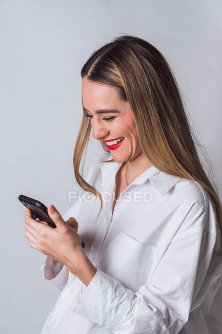 Positive junge schwangere Frau mit roten Lippen im weißen Hemd SMS auf Handy in der Nähe der grauen Wand — Stockfoto