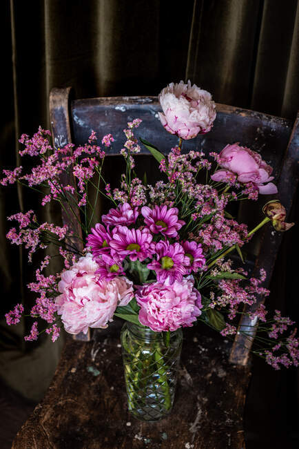 Букет свіжих різнокольорових півоній і хризантем у скляній вазі розміщений на обвітреному дерев'яному стільці біля штор у світлій кімнаті — стокове фото