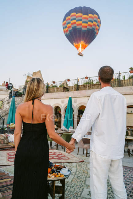 Back view of unrecognizable couple standing on the street cafe holding hands and watching hot air balloon flight — Stock Photo
