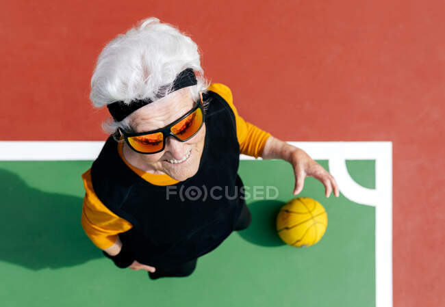 Зверху позитивно зріла жінка в спортивному одязі і сонцезахисних окулярах стоїть на спортивному майданчику з жовтим м'ячем під час гри в баскетбол — стокове фото