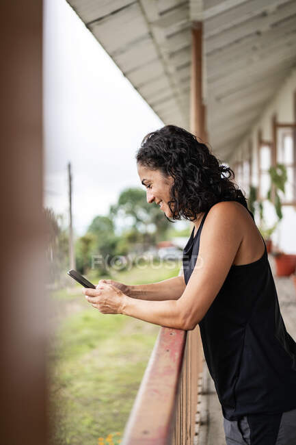 Vista lateral da jovem senhora étnica alegre com cabelos escuros encaracolados em roupas casuais sorrindo enquanto mensagens no telefone móvel em pé no terraço de madeira da casa envelhecida no campo — Fotografia de Stock