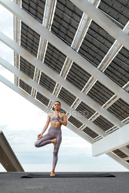Повний набір активних босоногих жінок у активаціях, що практикують позу Врікшасани, стоячи біля сонячної панелі під час тренування йоги в Барселоні. — стокове фото
