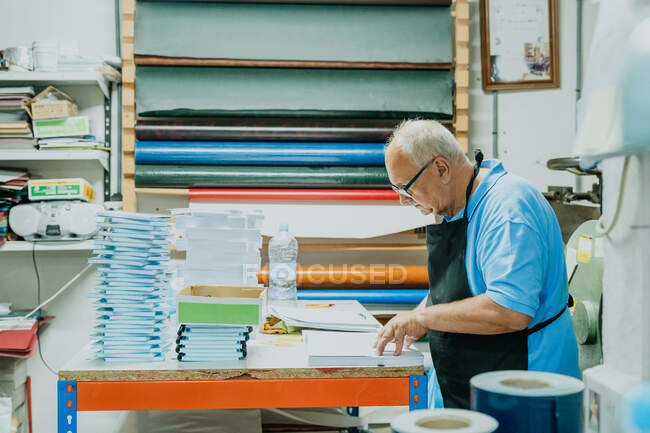 Vista lateral do artesão masculino sénior concentrado com cabelos brancos em avental e óculos em pé à mesa com pilha de papéis e caderno na oficina de impressão — Fotografia de Stock