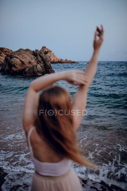 Vue arrière de la jeune femme attrayante dans des vêtements d'été élégants debout avec les bras levés dans l'eau de mer le soir — Photo de stock