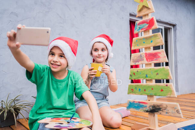 Optimistische Kinder beim Selbstporträt auf dem Smartphone, während sie bei der gemeinsamen Urlaubsvorbereitung zu Hause neben dem hölzernen Weihnachtsbaum sitzen — Stockfoto