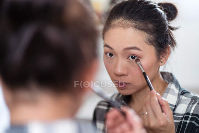 Очаровательная этническая женщина, нанося тени для век кистью, делает макияж и смотрит в зеркало — стоковое фото