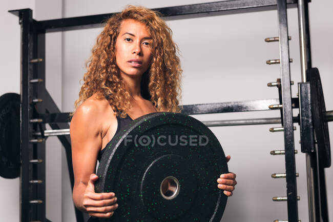 Femme de sport forte avec les cheveux bouclés en vêtements de sport debout et mettre la plaque de poids sur haltère pendant l'entraînement dans la salle de gym — Photo de stock