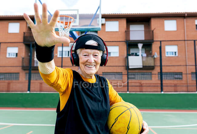 Усміхнена зріла жінка в навушниках з м'ячем в руці дивиться на камеру, стоячи на спортивному майданчику під час тренування з баскетболу — стокове фото