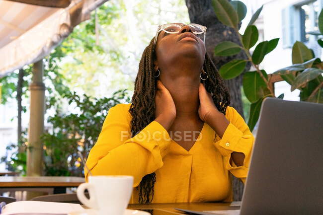 Thoughtful African American female freelancer in eyeglasses looking up while sitting at table with netbook and cup of hot drink in cafeteria — Stock Photo