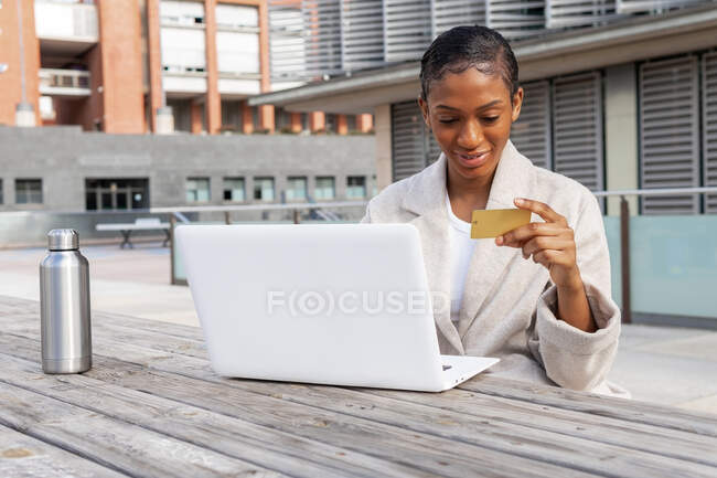 Femme avec carte de crédit à la main tapant sur le netbook moderne tout en faisant des achats en ligne dans la rue en ville — Photo de stock