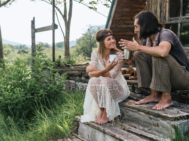Pareja hippie descalza compartiendo un café mientras hablan y se miran en un porche de madera - foto de stock