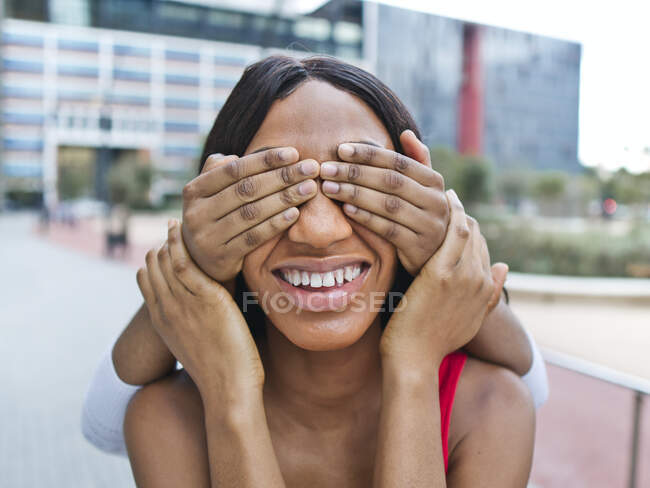 Беззернистый человек, закрывающий глаза улыбающейся афроамериканки руками, стоя на улице со зданием в городе — стоковое фото