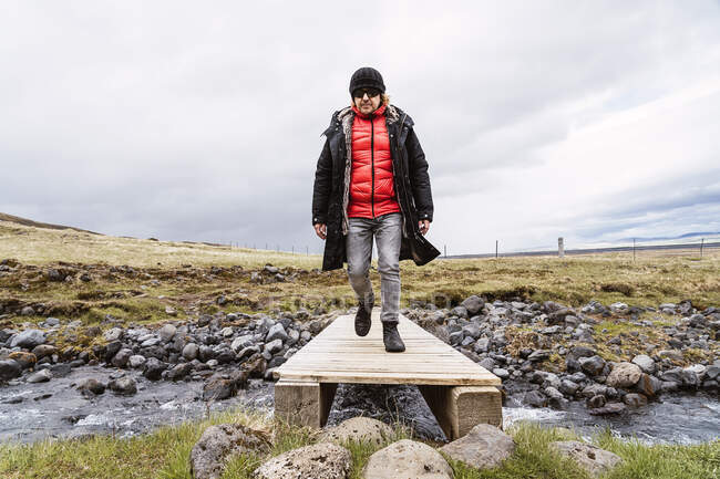 Full body of young male traveler in warm outerwear hat and sunglasses walking on small wooden bridge crossing wild river during trip in Iceland on cloudy day — Stock Photo