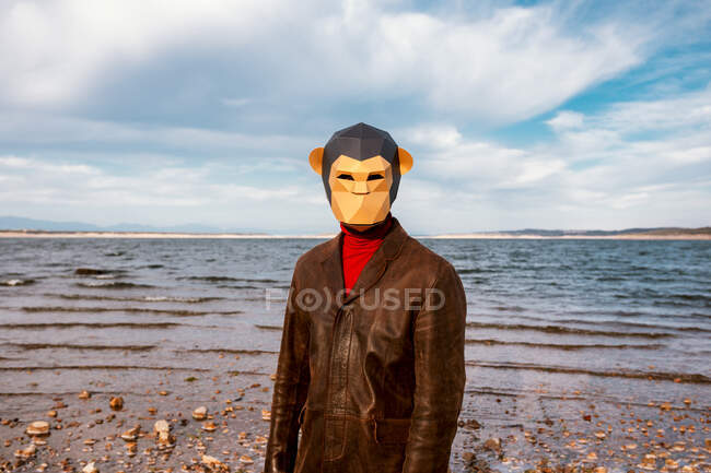 Persona irriconoscibile in giacca marrone e maschera geometrica scimmia in piedi su sfondo di mare increspato in estate — Foto stock