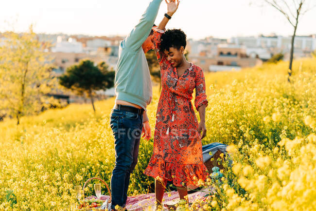 Glückliches junges, vielfältiges Paar Händchen haltend beim gemeinsamen Tanzen auf blühender Wiese beim romantischen Picknick am sonnigen Tag — Stockfoto
