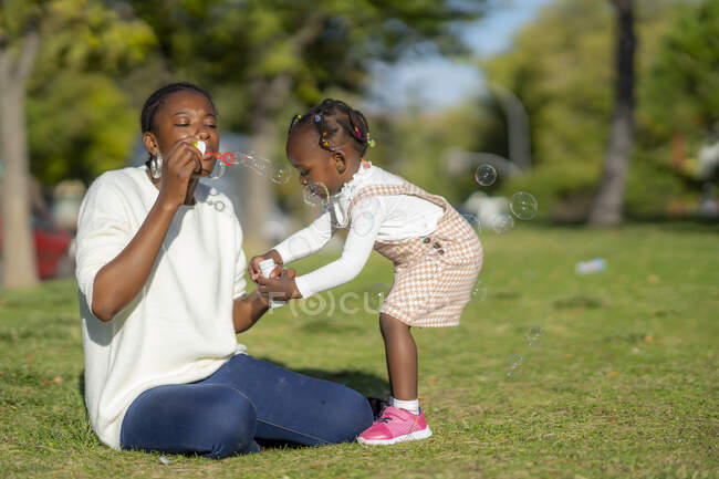 Calmo mulher afro-americana sentada na grama verde e soprando bolhas de sabão enquanto brincava com a filha no parque no horário de verão — Fotografia de Stock