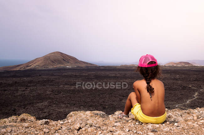 Indietro vista di anonimo bambino senza maglietta che ammira la lava vulcanica e il mare mentre è seduto a Lanzarote Isole Canarie Spagna — Foto stock