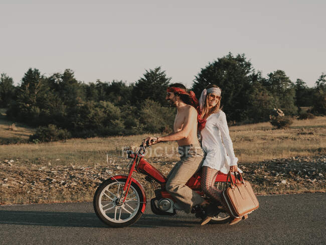 Side view of full body of hippie couple riding on red moped on asphalt roadway during trip in nature with trees on summer day — Stock Photo