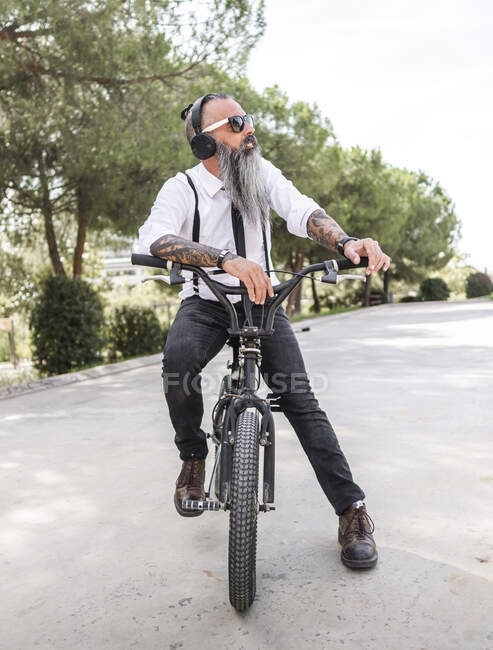 Confiado hipster masculino en camisa blanca escuchando música en auriculares mientras está sentado en bicicleta en la calle con árboles verdes - foto de stock