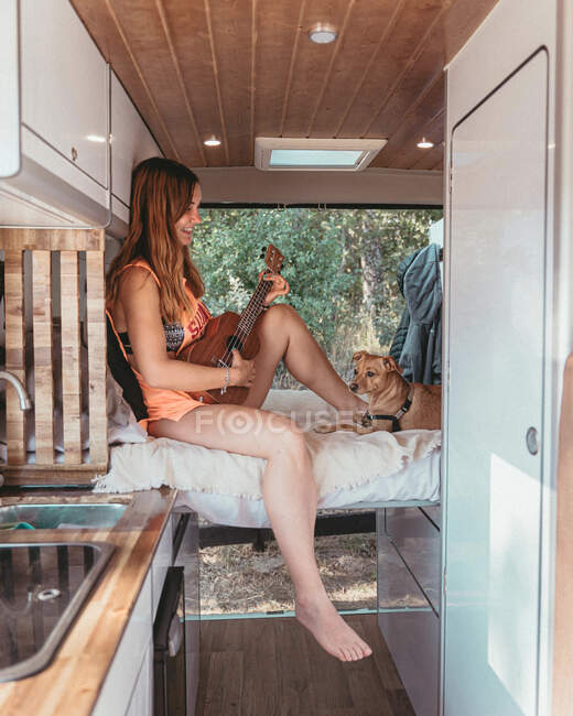 Side view of cheerful female traveler sitting on bed with dog in van and playing ukulele during trip in Spain — Stock Photo
