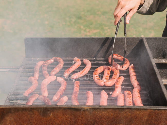 Cropped unrecognizable chef with tongs cooking various types of tasty sausages roasting on grill grate above charcoal in countryside during barbecue in countryside on summer day — Stock Photo