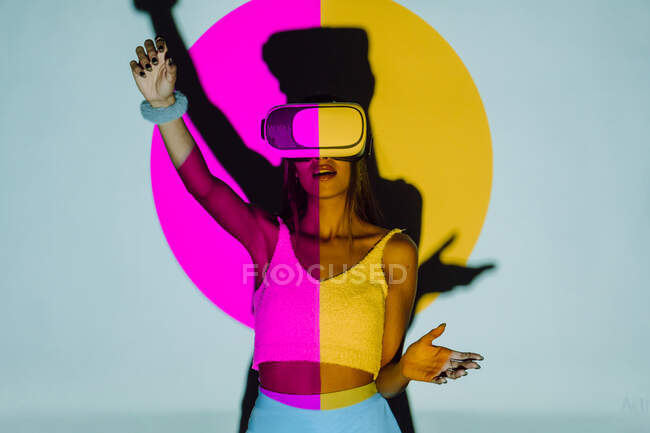 Femme surprise avec bras levé explorant la réalité virtuelle dans un casque tout en se tenant debout dans la lumière du projecteur rose et jaune sur fond gris — Photo de stock