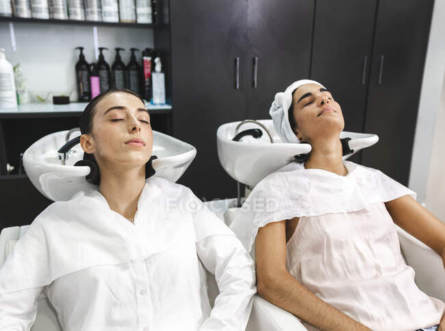 Jeunes femmes en vêtements blancs fermant les yeux tout en ayant les cheveux lavés dans un salon de beauté moderne — Photo de stock