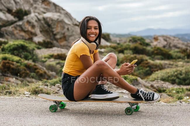 Corpo pieno di femmina etnica positiva con cuffie wireless sul collo che navigano sullo smartphone mentre siede sul longboard sulla strada asfaltata a guardare la fotocamera — Foto stock