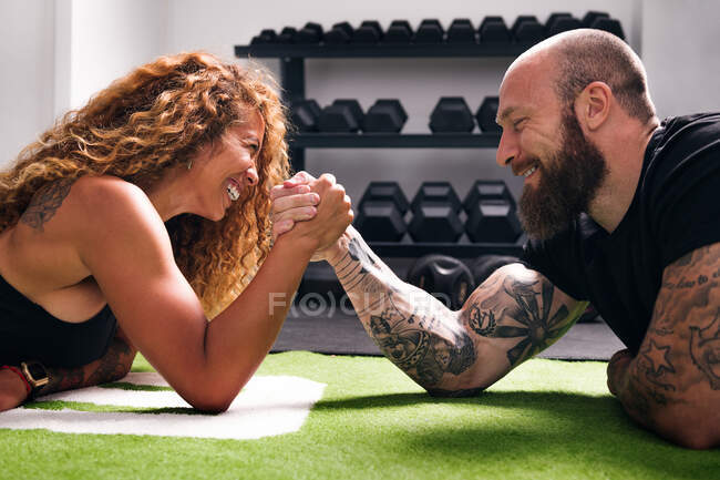 Side view of smiling hispanic sportswoman with curly hair holding hand of cheerful bodybuilder during arm wrestling challenge against dumbbells in gym — Stock Photo