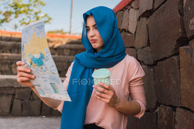 Сконцентрована мусульманка в хустці і з витягуванням кави на паперовій мапі і в пошуках напрямку стоячи біля кам'яної стіни. — стокове фото