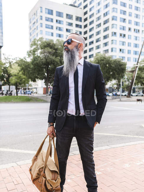 Hombre hipster barbudo de confianza en traje elegante con bolsa de pie en la acera cerca de la carretera en la calle con edificios en la ciudad - foto de stock