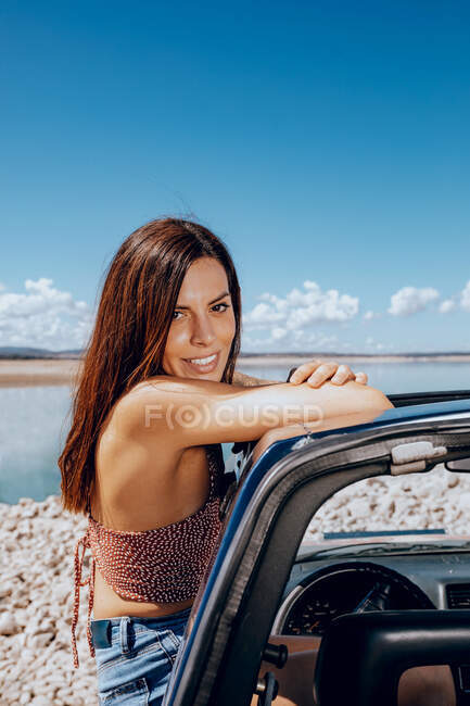 Side view of young anonymous female in denim jeans and top leaning out of car window looking at camera on coast of pond — Stock Photo